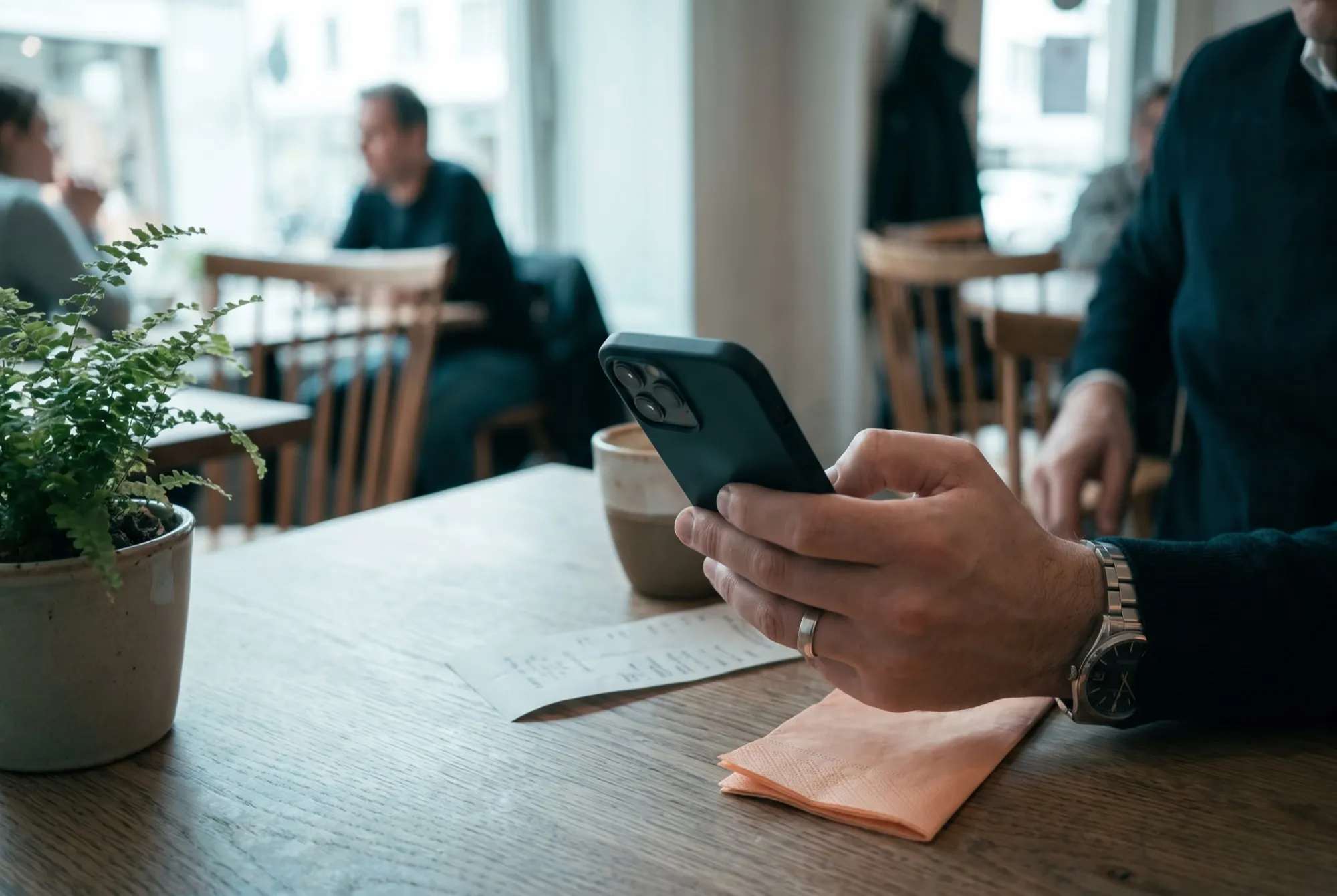 Eine Hand hält im Café ein Smartphone über eine Quittung, daneben Kaffeetasse und Pflanze