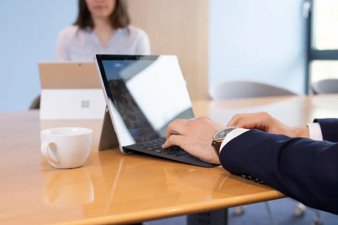 Hände auf der Tastatur eines Tablets, daneben eine Kaffeetasse, im Hintergrund eine Kollegin