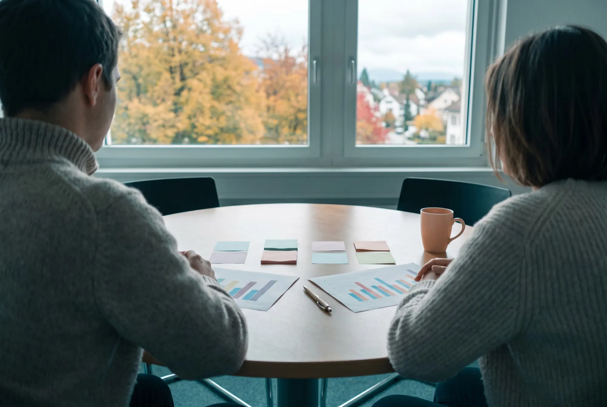 Besprechungstisch im Herbst, zwei Personen von hinten vor Diagrammen und Notizzetteln, draussen Herbstbäume