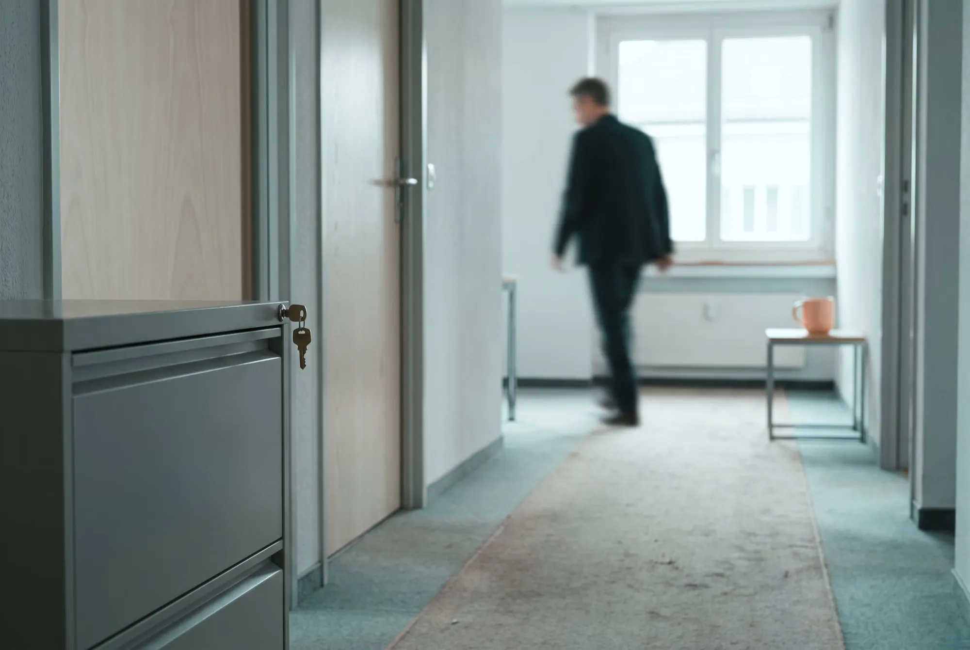 Büroflur mit abgeschlossenem Aktenschrank, der Schlüssel steckt, im Hintergrund eine Person unscharf
