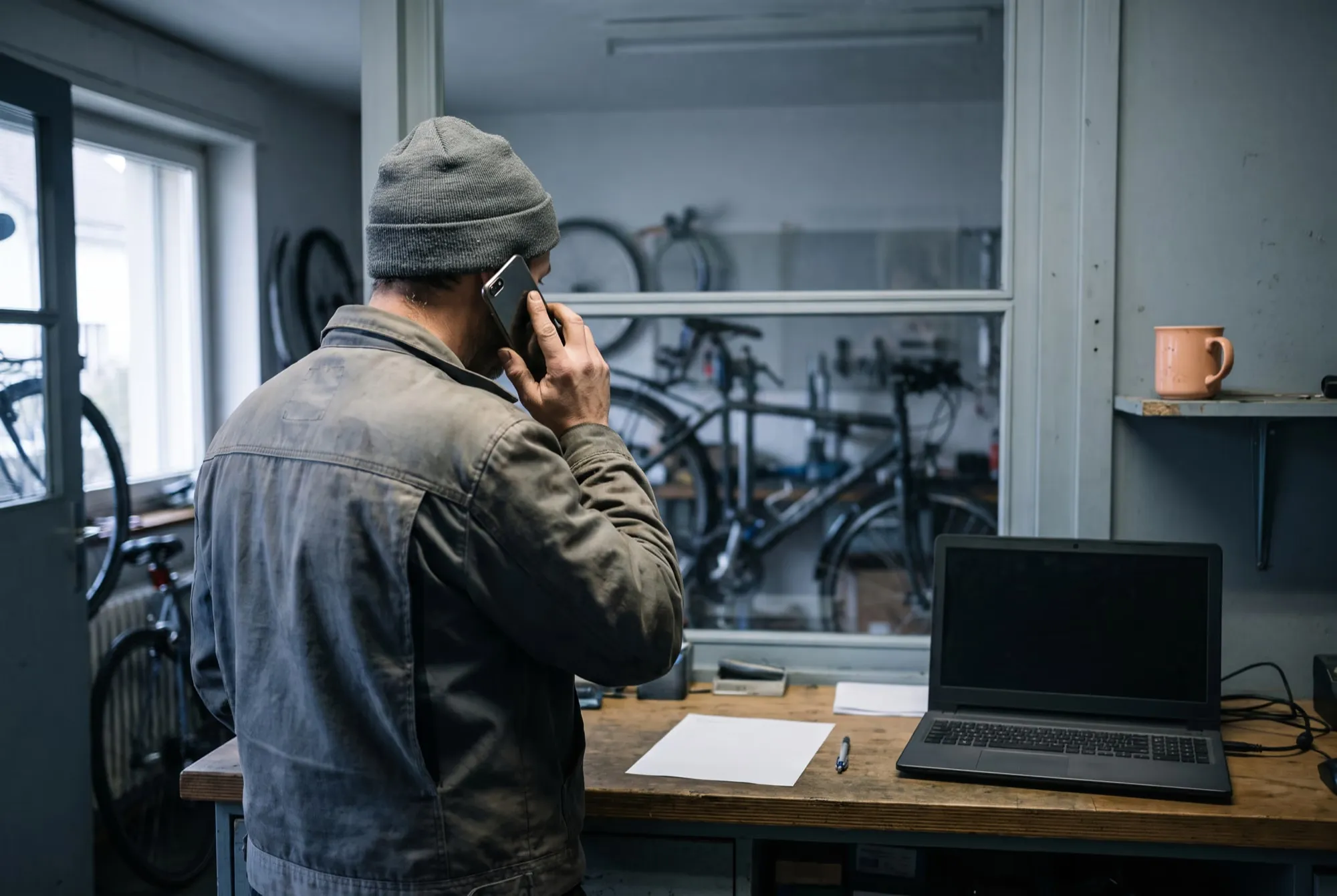 Inhaber einer Velowerkstatt telefoniert am Werkstatttresen, daneben ein Laptop und ein leeres Blatt Papier