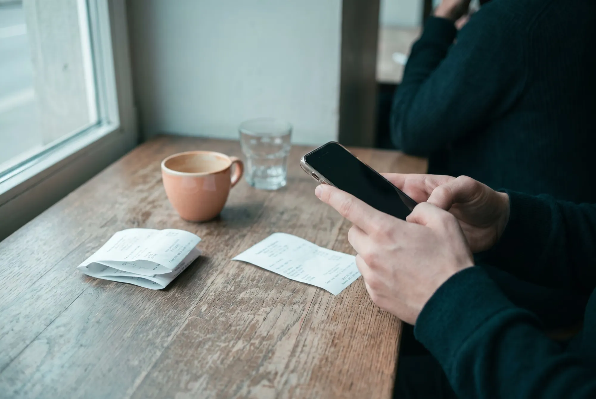 Hände fotografieren mit dem Smartphone eine Quittung auf einem Holztisch, daneben eine Kaffeetasse