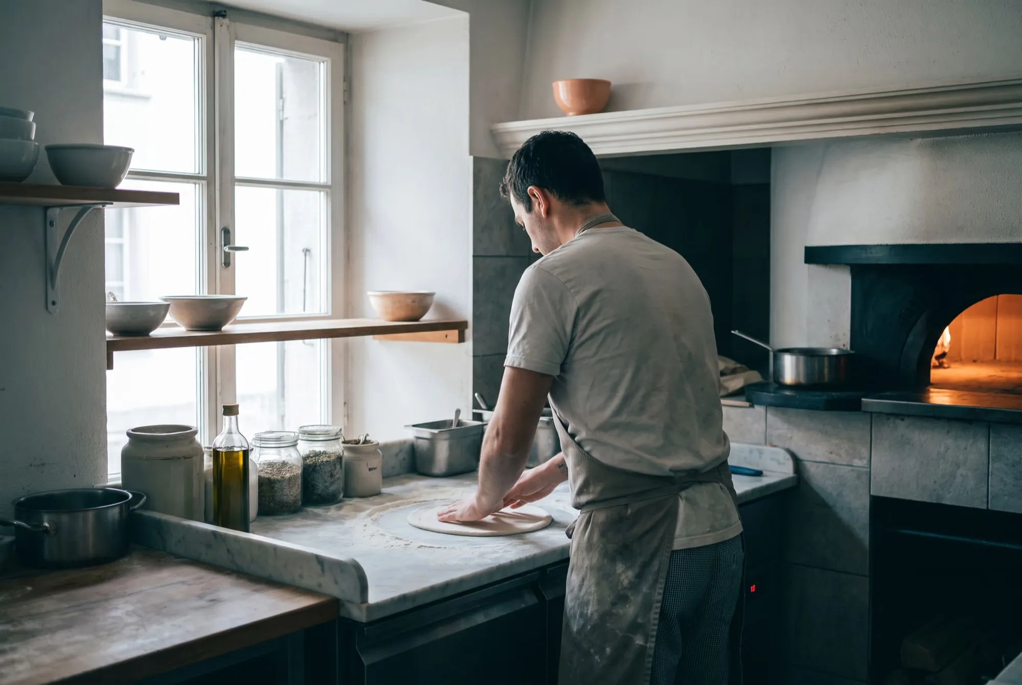 Pizzeria-Küche mit Holzofen, ein Pizzaiolo zieht auf der bemehlten Marmorplatte einen Teig aus