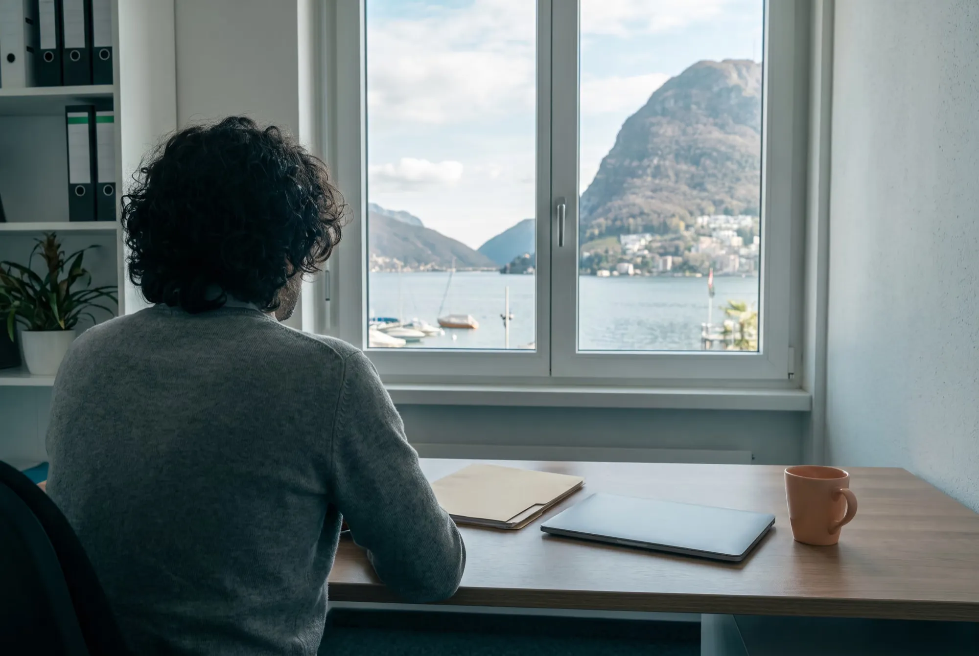 Büro im Tessin, eine Person von hinten am Schreibtisch, durchs Fenster der Luganersee