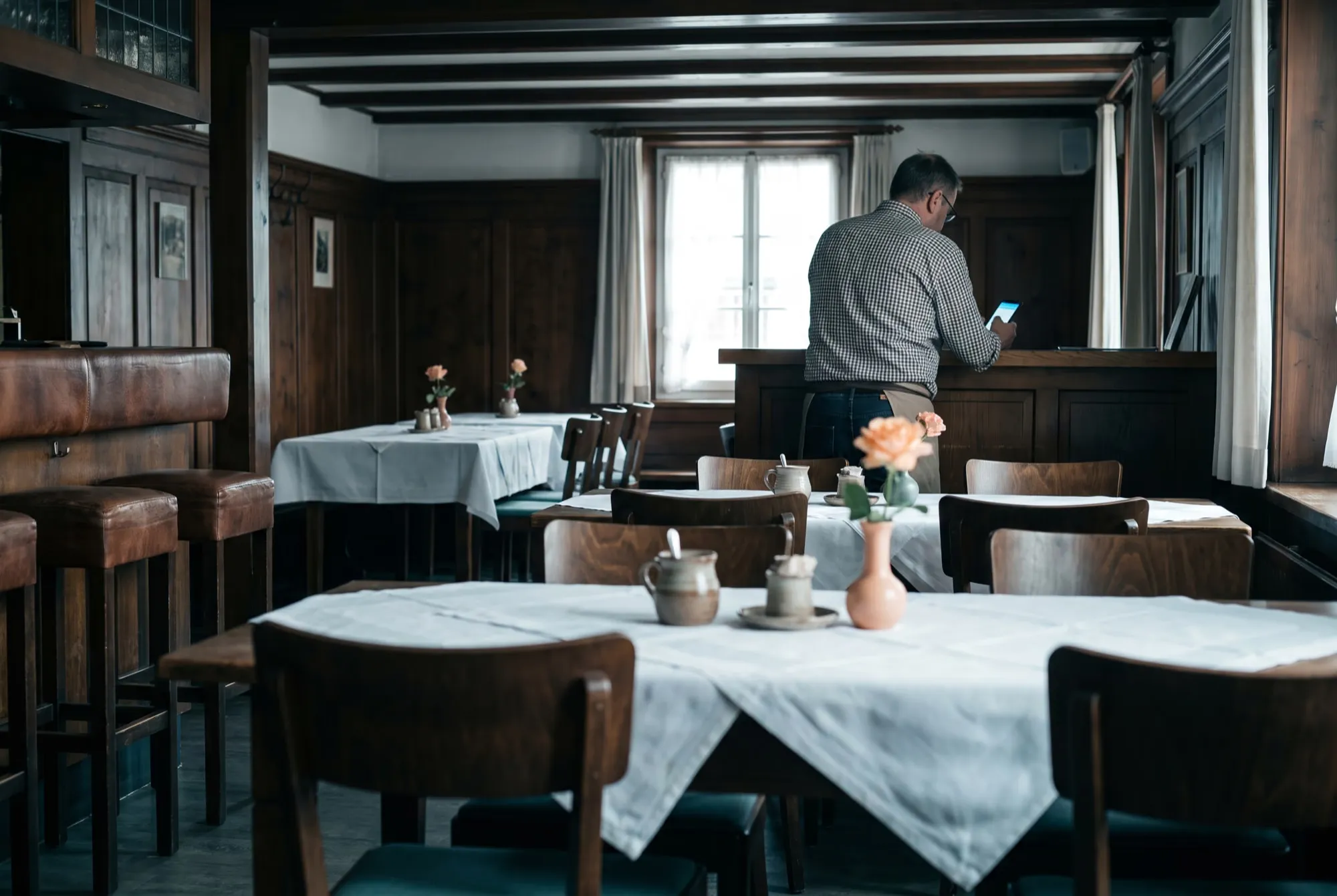 Gaststube am Morgen vor dem Service, gedeckte Tische, ein Gastgeber von hinten am Buffet mit dem Smartphone