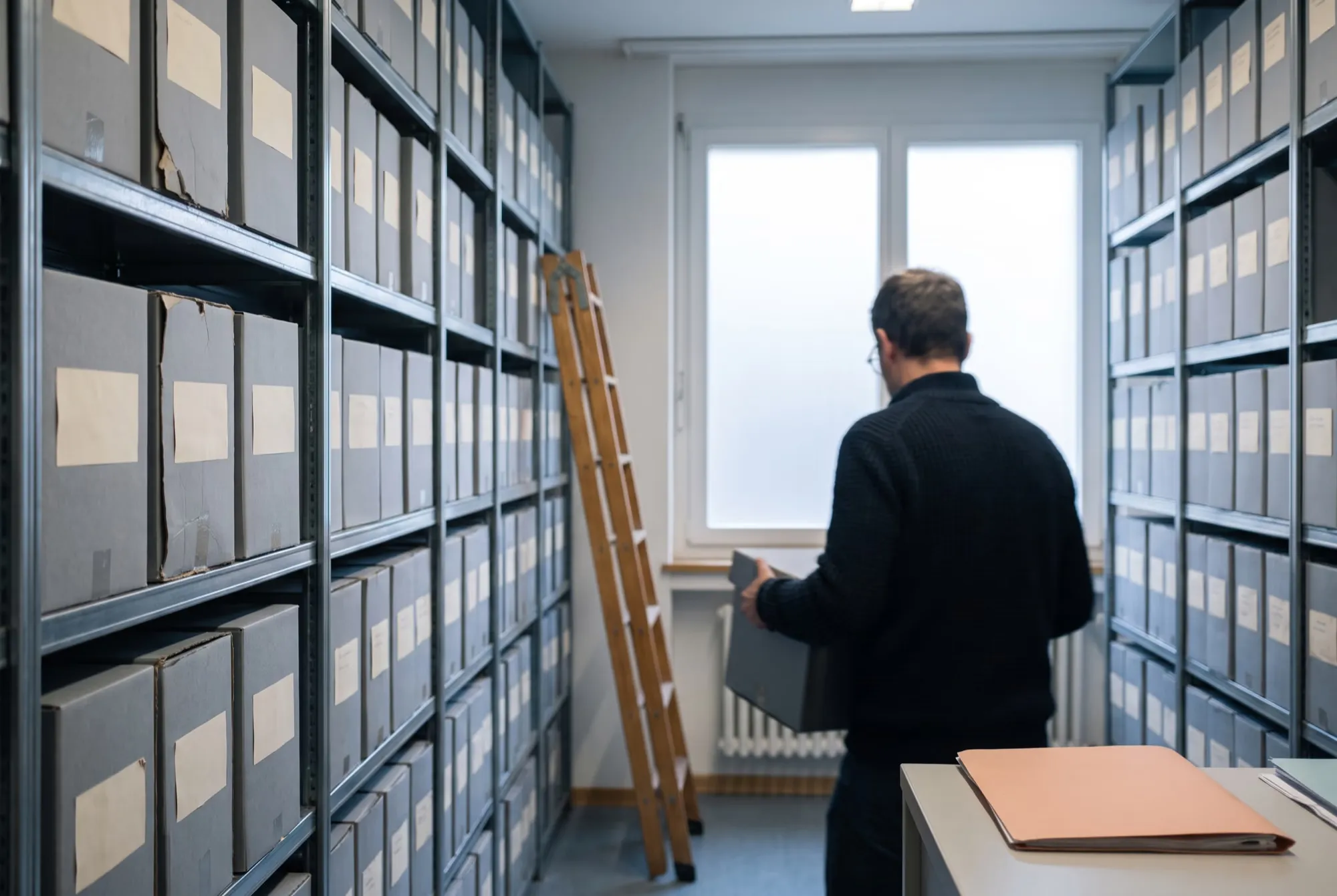 Archivraum mit Regalen voller grauer Archivschachteln, ein Mitarbeiter von hinten mit einer Schachtel